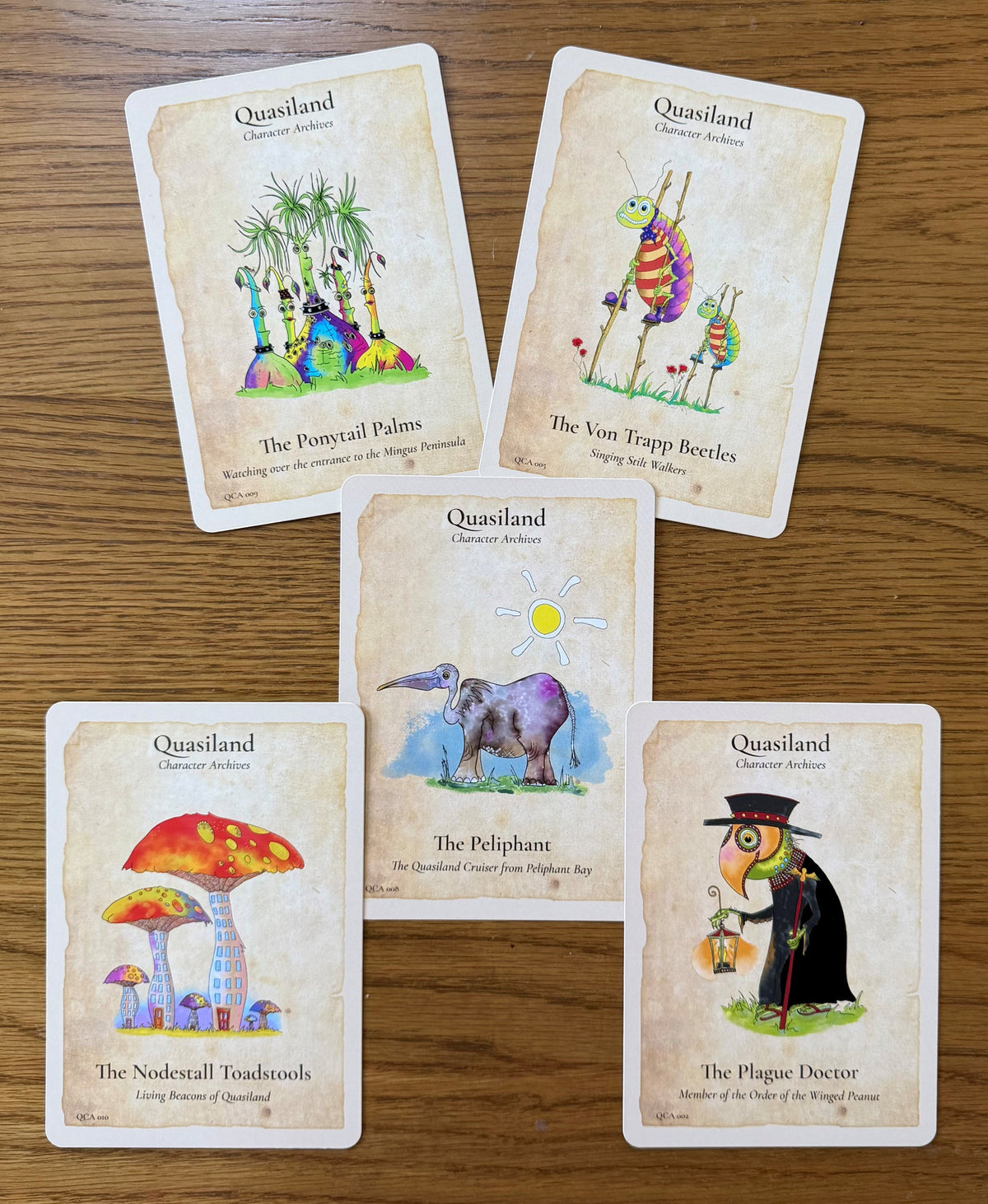 Five illustrated collector cards from the Quasiland deck arranged on a wooden tabletop. The cards feature whimsical artwork and titles including The Ponytail Palms, The Von Trapp Beetles, The Peliphant, The Nodestall Toadstools, and The Plague Doctor.